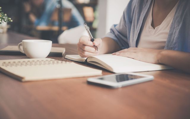 Personne notant une idée de recette dans un carnet, à côté d'une tasse et d'une tablette